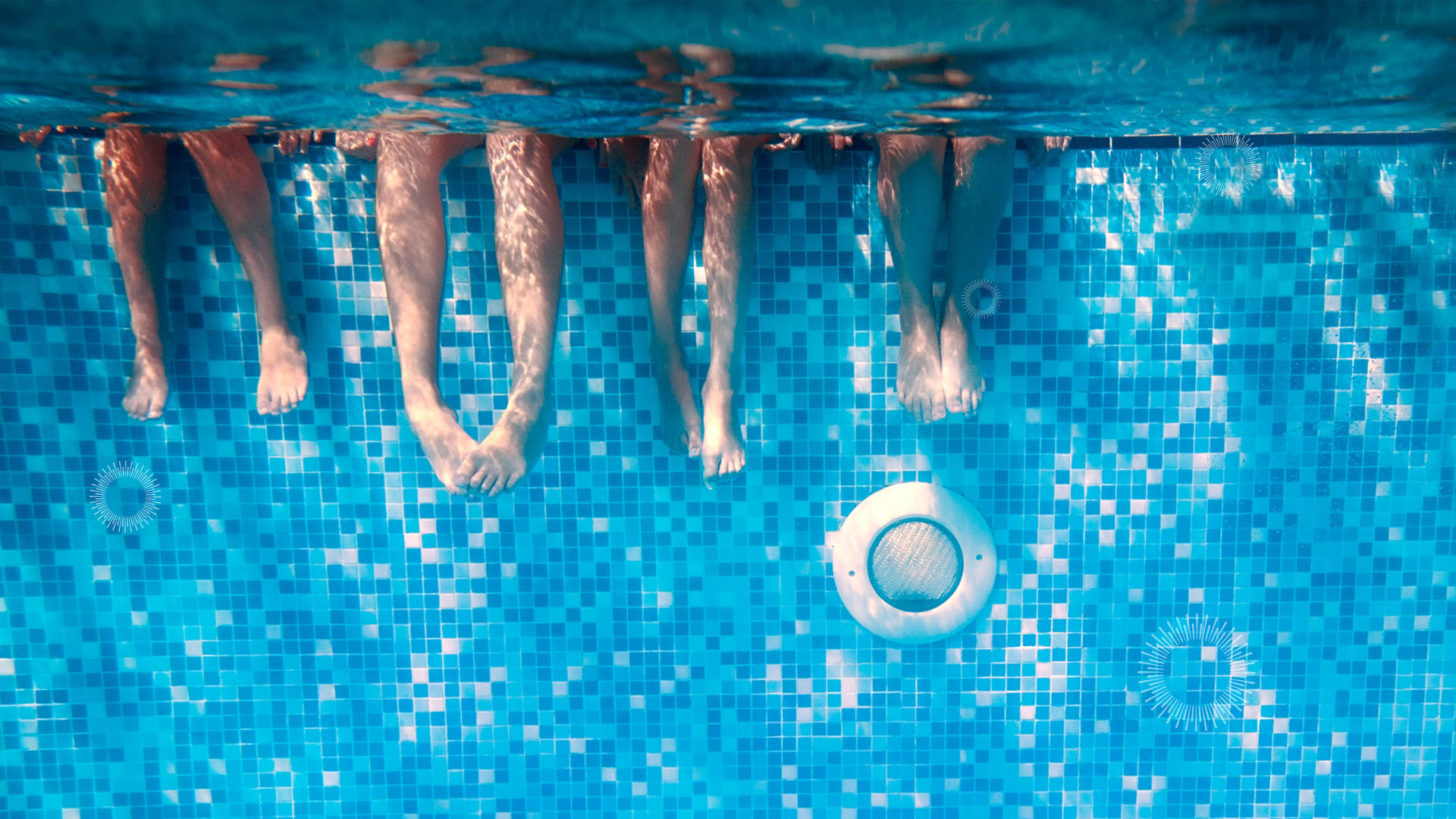 underwater view of the legs of four kids sitting on the edge of a pool with blue tile in the background