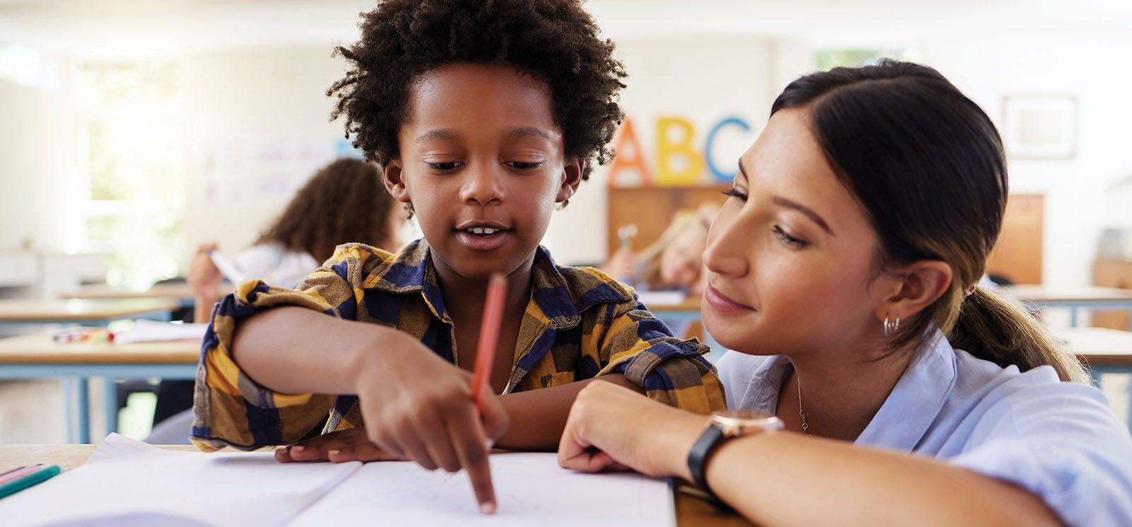 young boy holding pen and pointing to notebook page while teacher looks on