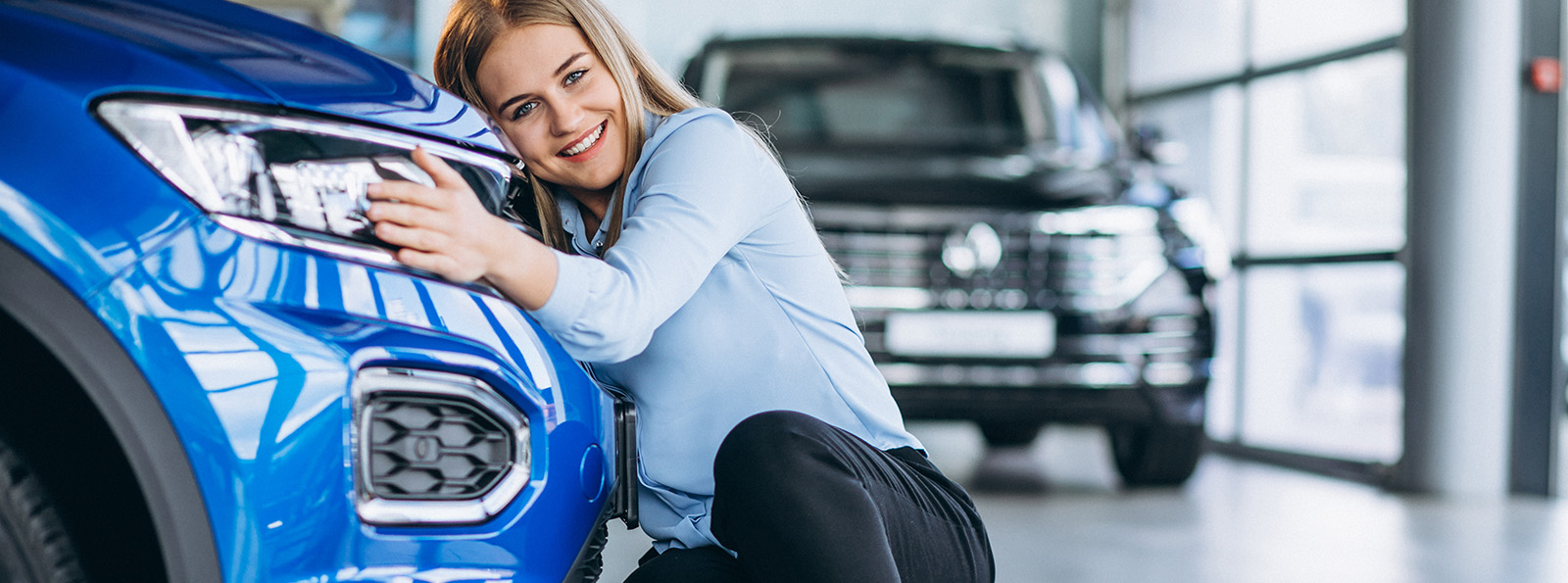 A young woman in a car showroom smiles while crouching to hug the front of a new blue car
